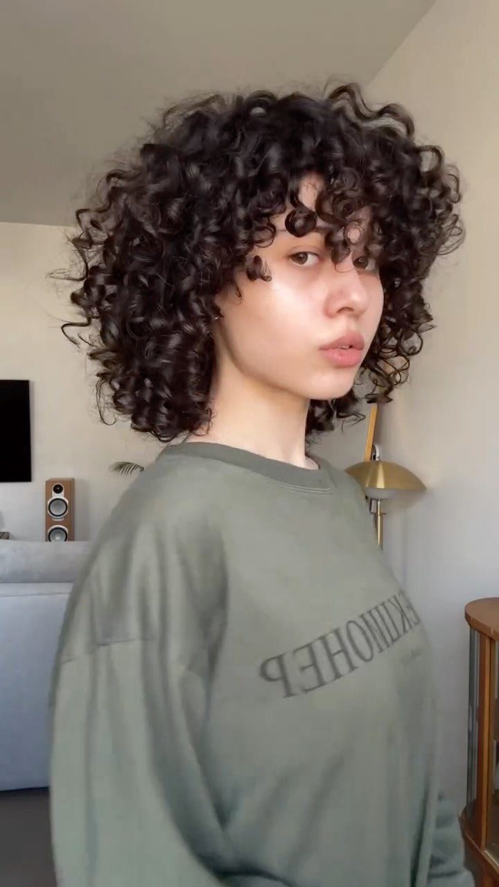 2C Curly Hairstyles: 10 Ideas for Layers, Buns and Bangs 10 Person with voluminous dark brown curly hair wearing an olive green sweatshirt indoors.