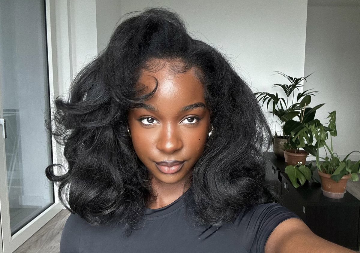 4C Hair Blown Out with Braids, Buns and Waves: 9 Ideas 9 Woman with voluminous, wavy blown-out 4C hair posing indoors near green plants.