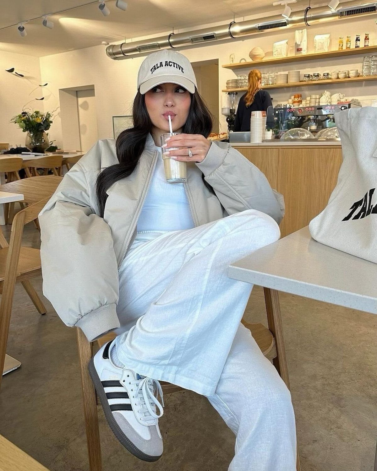 Woman in a cafe wearing grey Adidas Samba sneakers and a bomber jacket.
