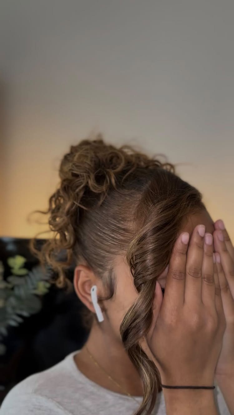High curly bun with a face-framing tendril on a person covering their face.