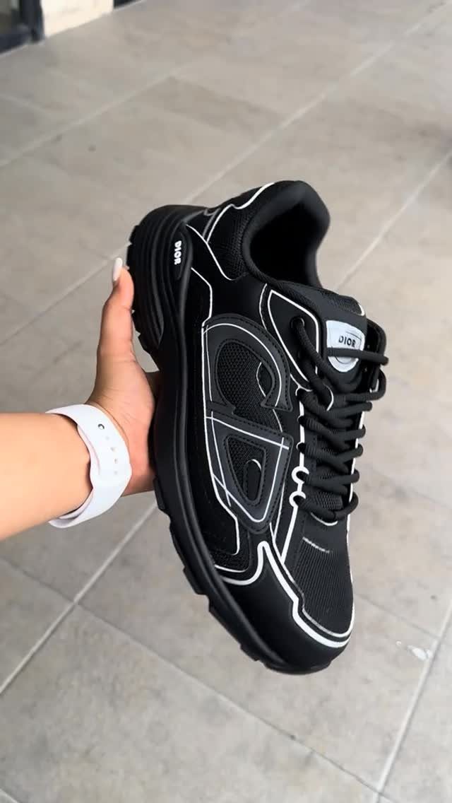 Black Dior sneaker with white accents held by a hand over grey tiles