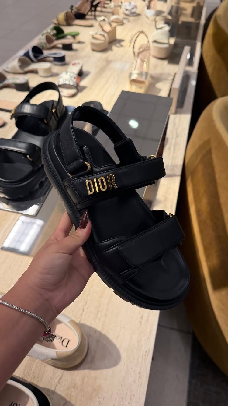 Black leather Dior slide sandal with gold logo held by a hand.