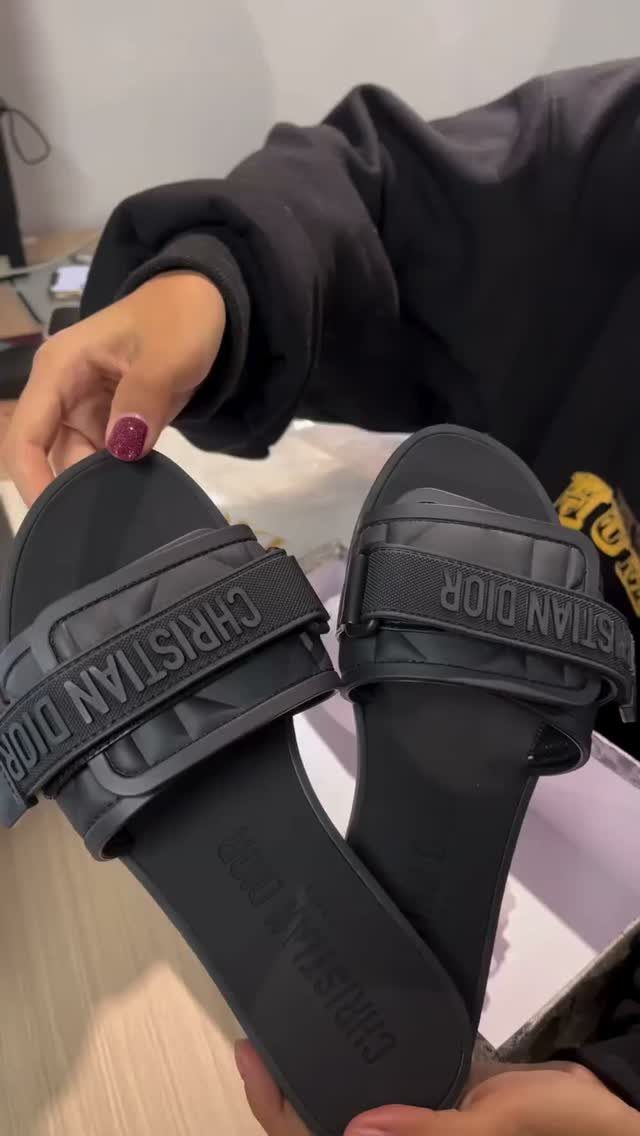 Black Dior slide sandals with embossed lettering held by a person in black.