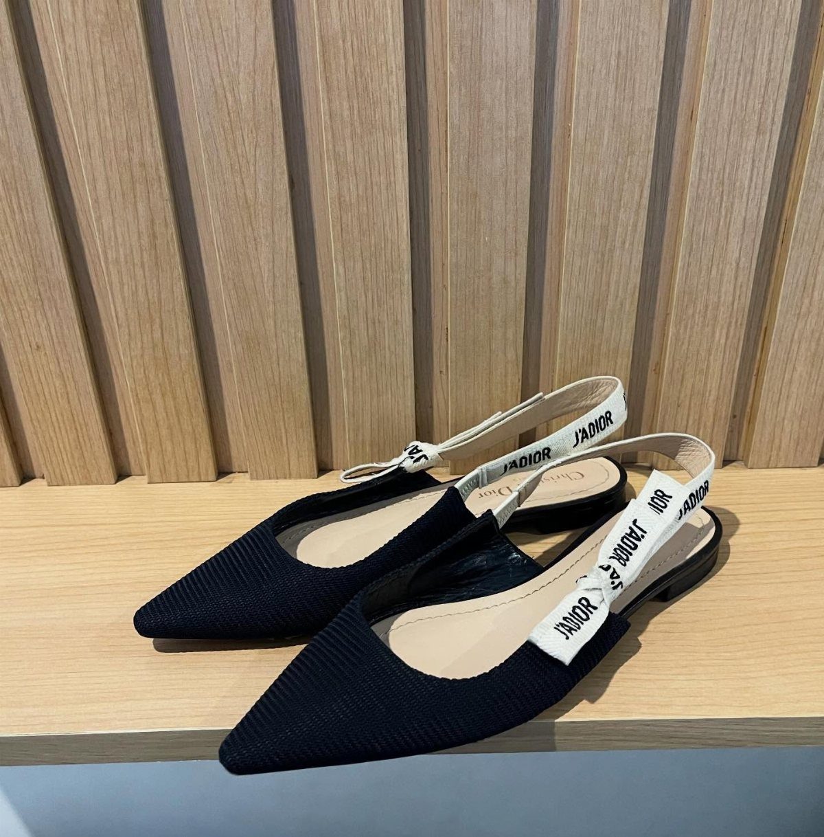 Black pointed-toe slingback flats with white J'Adior embroidered straps on a wooden shelf.