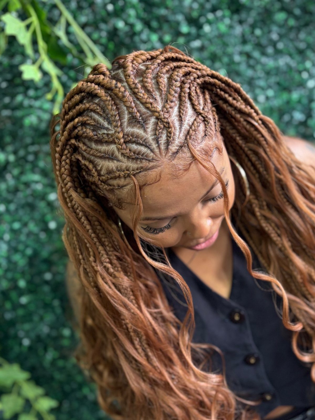 27 Boho Fulani Braids Hairstyles for Every Length and Look 24 Woman with reddish-brown boho Fulani braids and curly extensions against green foliage.