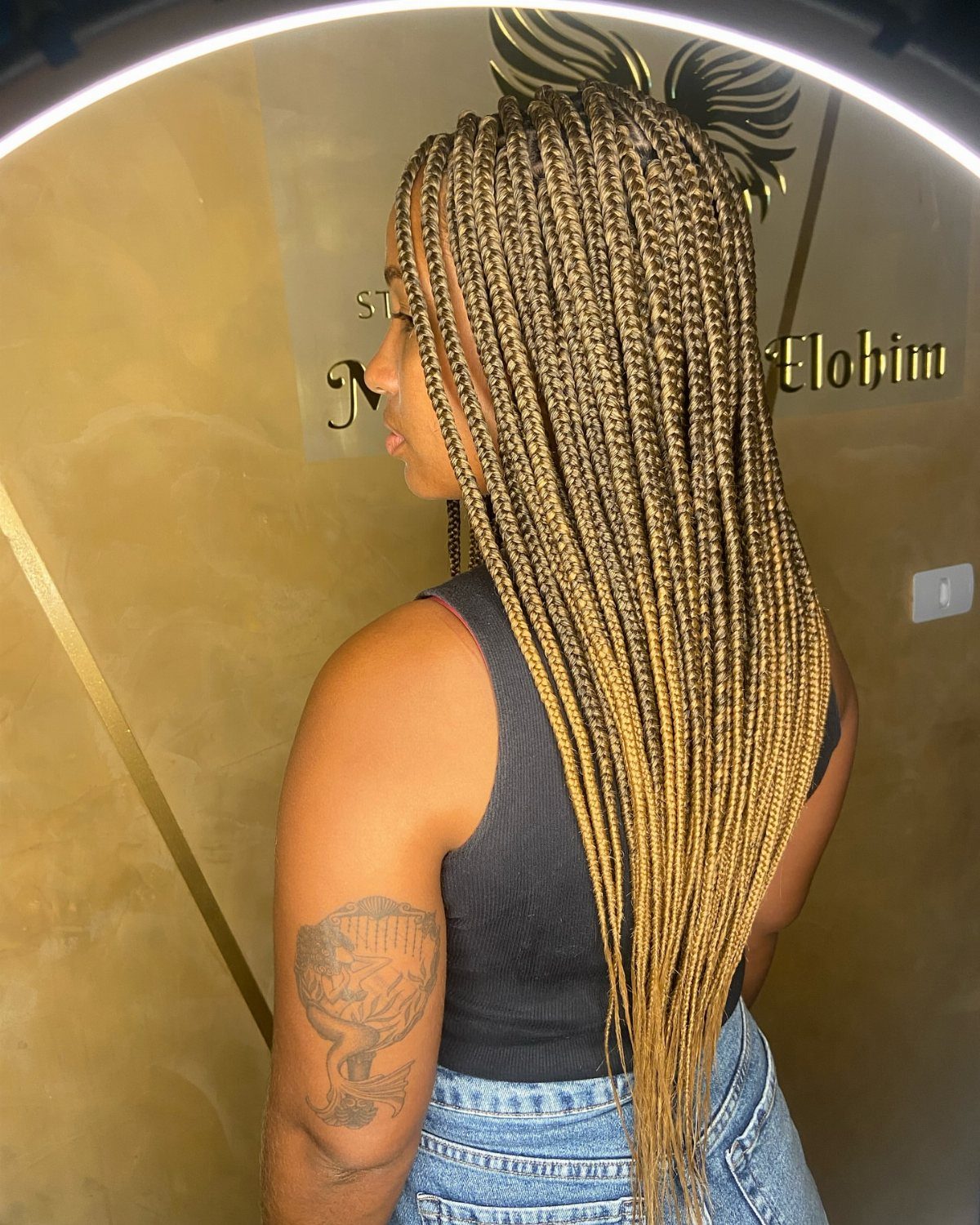 Woman with long honey-blonde box braids wearing a black top and blue jeans.