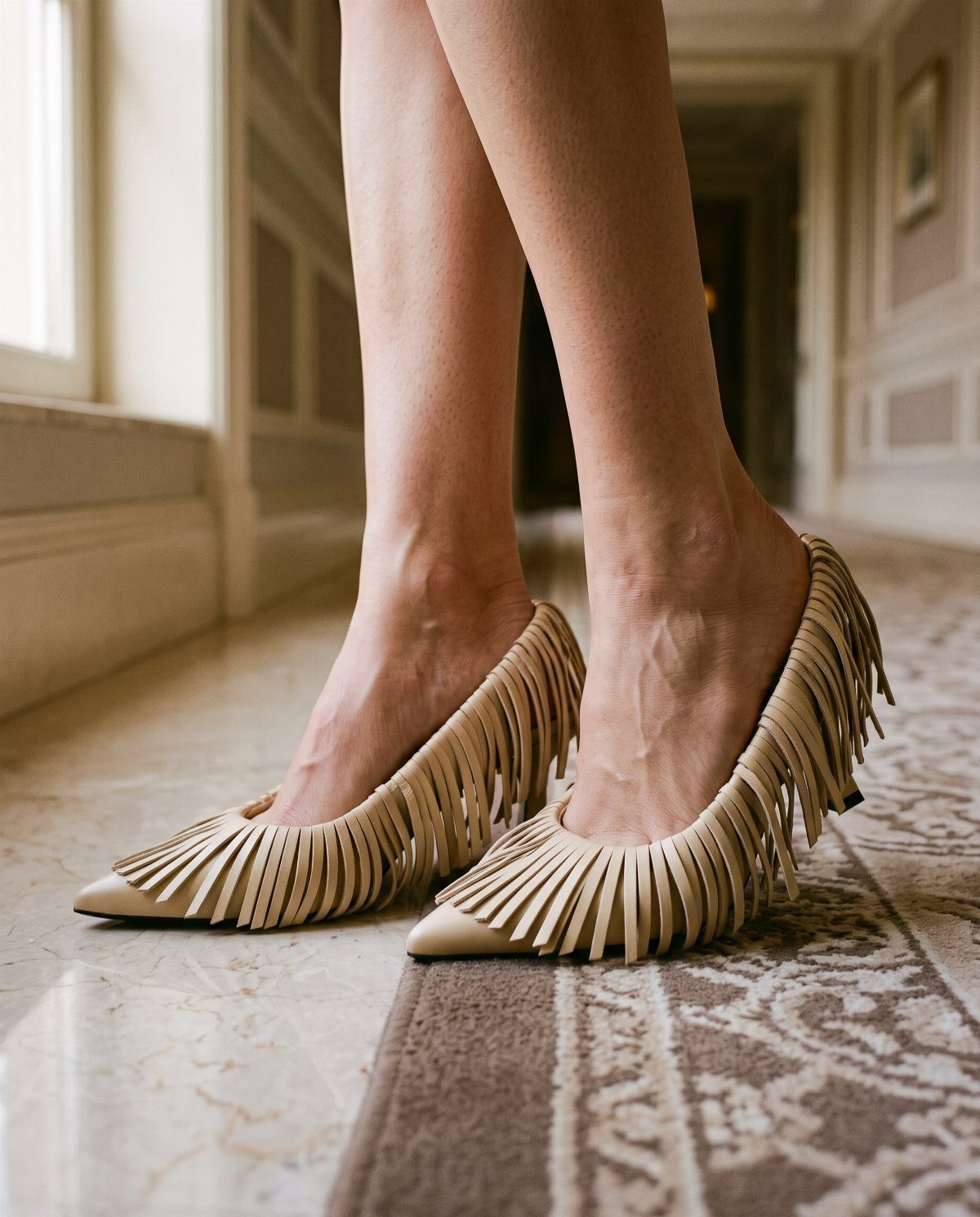 Tan pointed-toe heels with long fringe worn by a person on a rug.