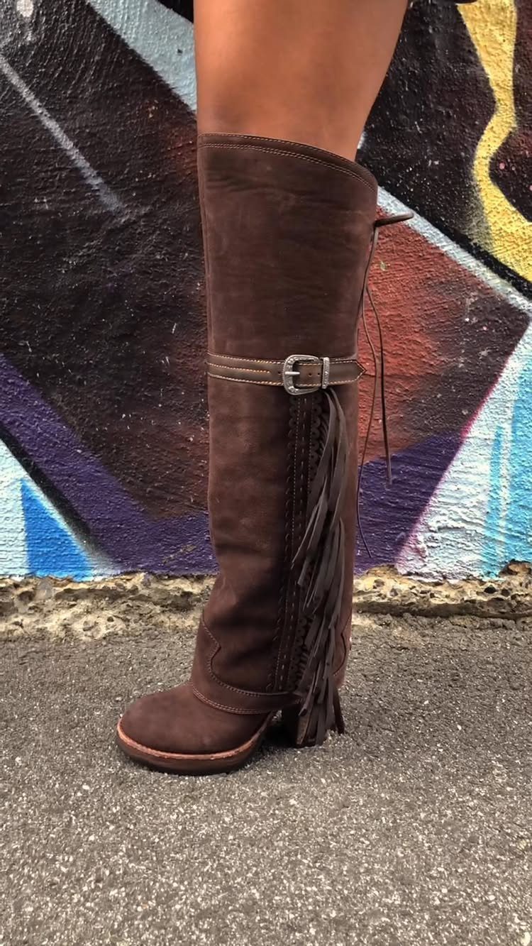 Brown suede knee-high boots with fringe and a buckle on a person.