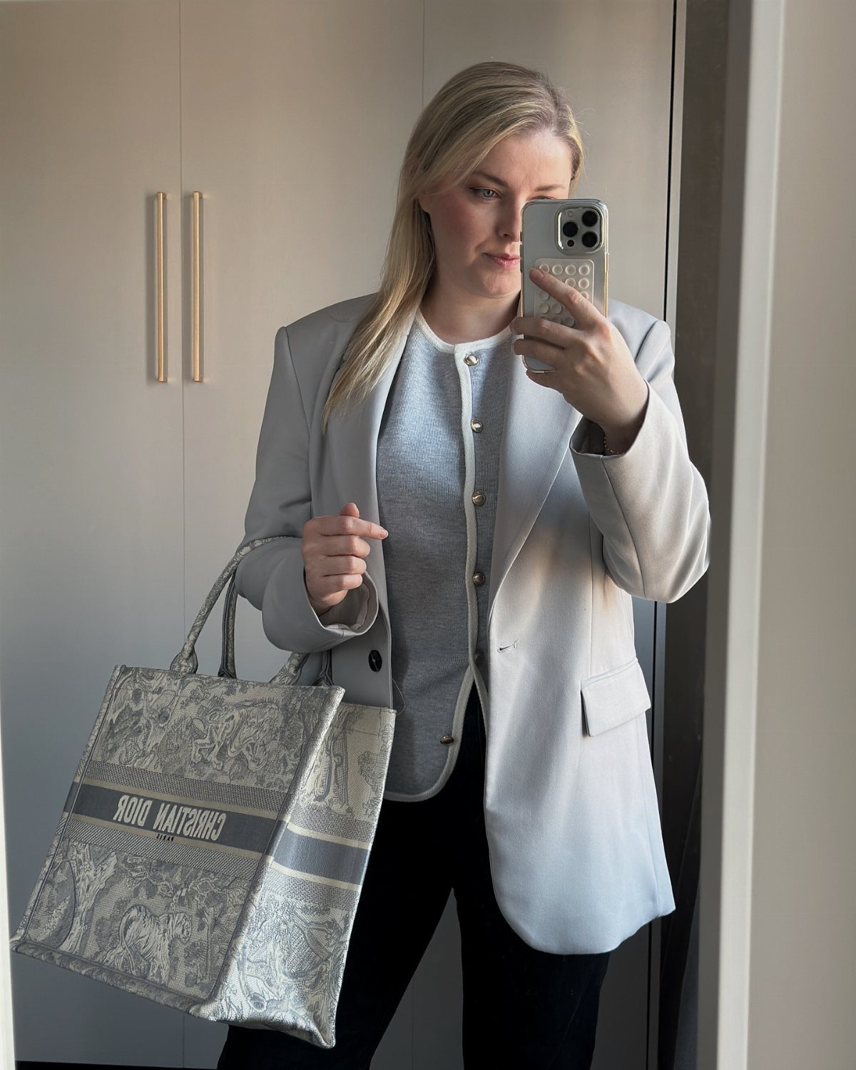 Woman in grey blazer and blue cardigan holding a Dior tote bag.