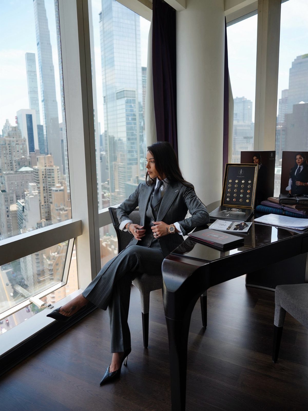Woman in a grey tailored business suit sitting at a desk in a skyscraper office.