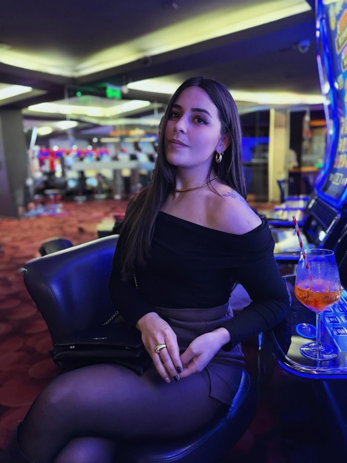 Woman in black off-the-shoulder top and grey skirt sitting in a casino lounge.