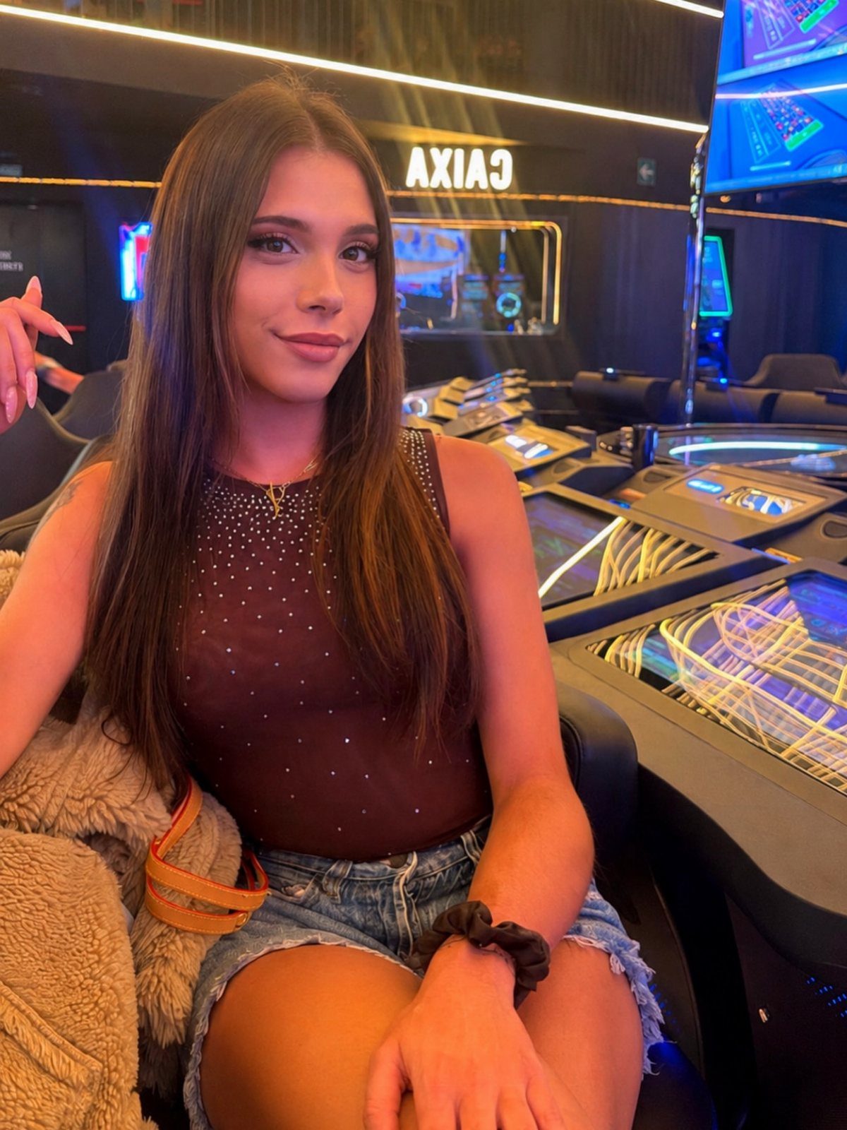 Woman in sparkling maroon top and denim shorts sitting at casino slot machines.