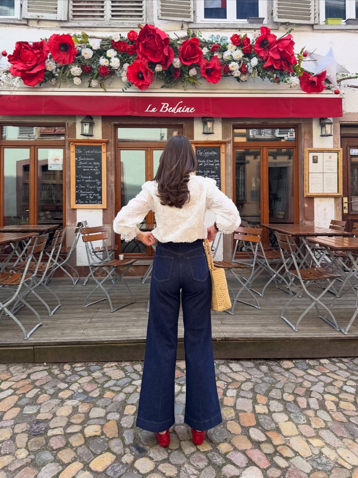 12 Casual Elegance Outfits for Easy Everyday Style 5 Woman in white blouse and wide-leg jeans standing before a flower-decorated cafe.