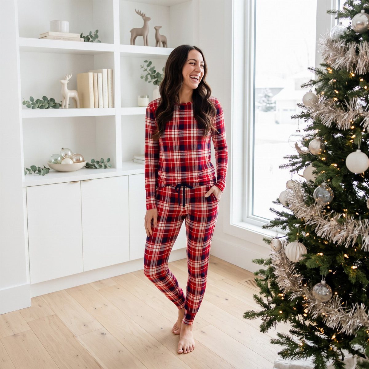 Woman in red plaid pajamas standing next to a decorated Christmas tree indoors.
