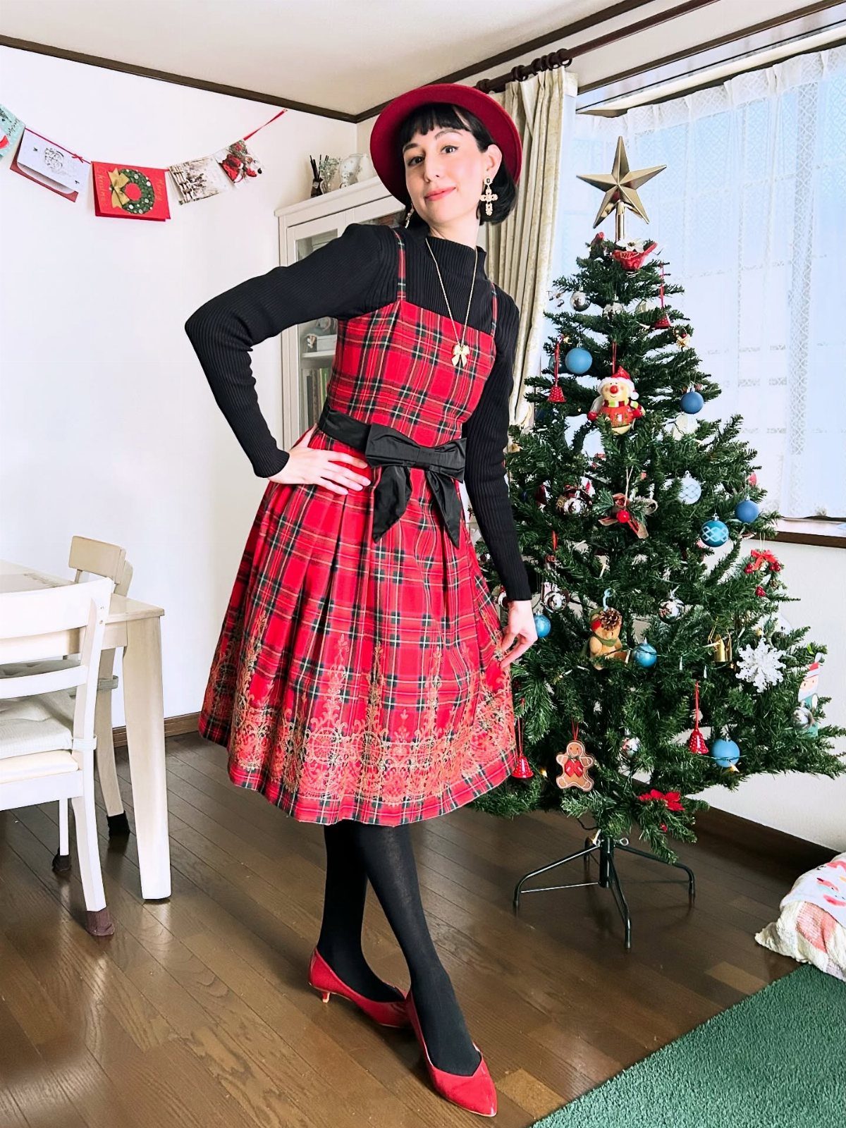 Woman in red plaid dress and black knit top standing by Christmas tree