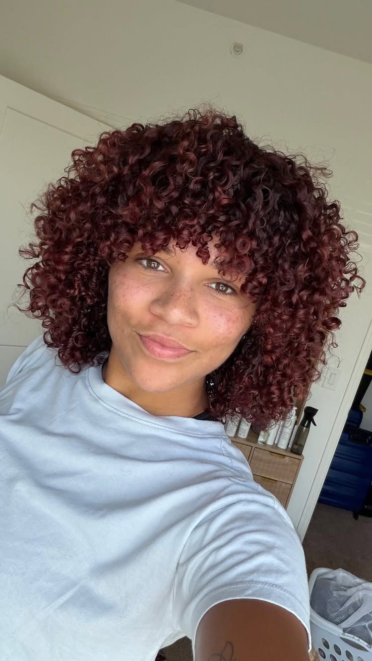 Person with voluminous curly reddish-brown hair and bangs wearing a white t-shirt.