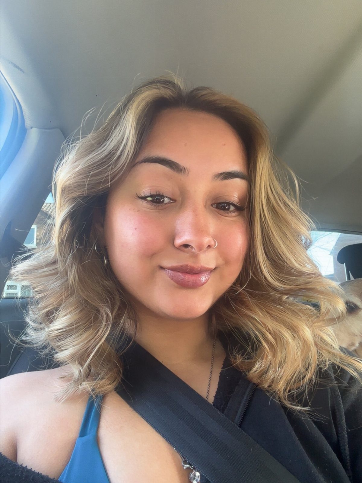 Woman with blonde-highlighted wavy hair posing inside a car with natural lighting.