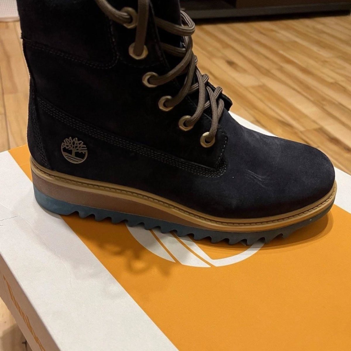 Dark blue suede boot with gold laces resting on a yellow shoe box.