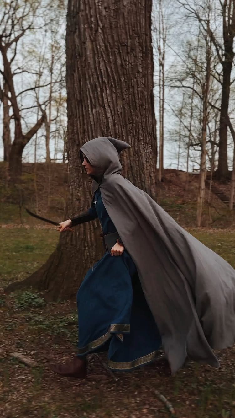 Person in a blue dress and grey hooded cloak holding a blade in woods