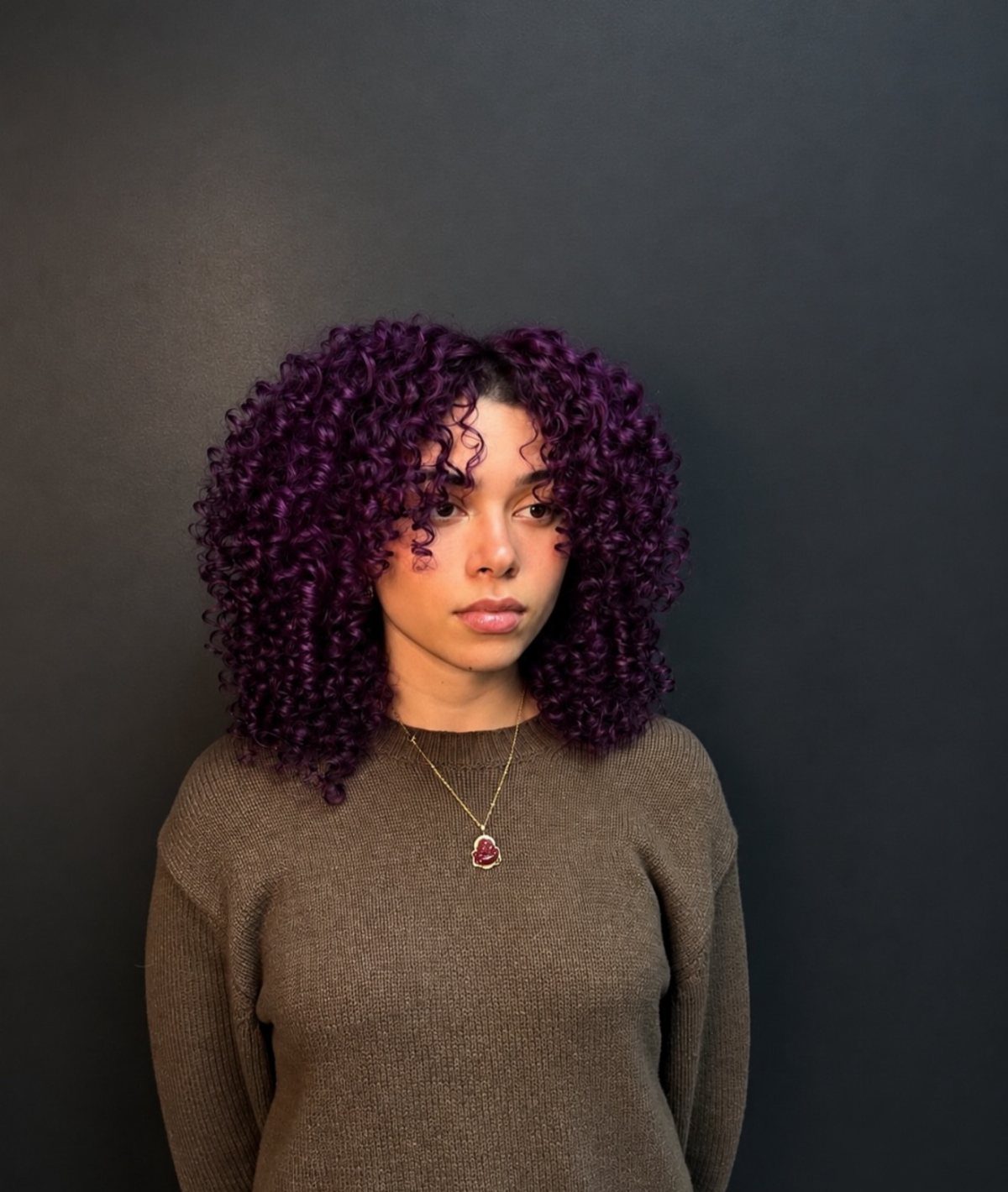 Woman with curly dark violet hair wearing a brown sweater and gold necklace.