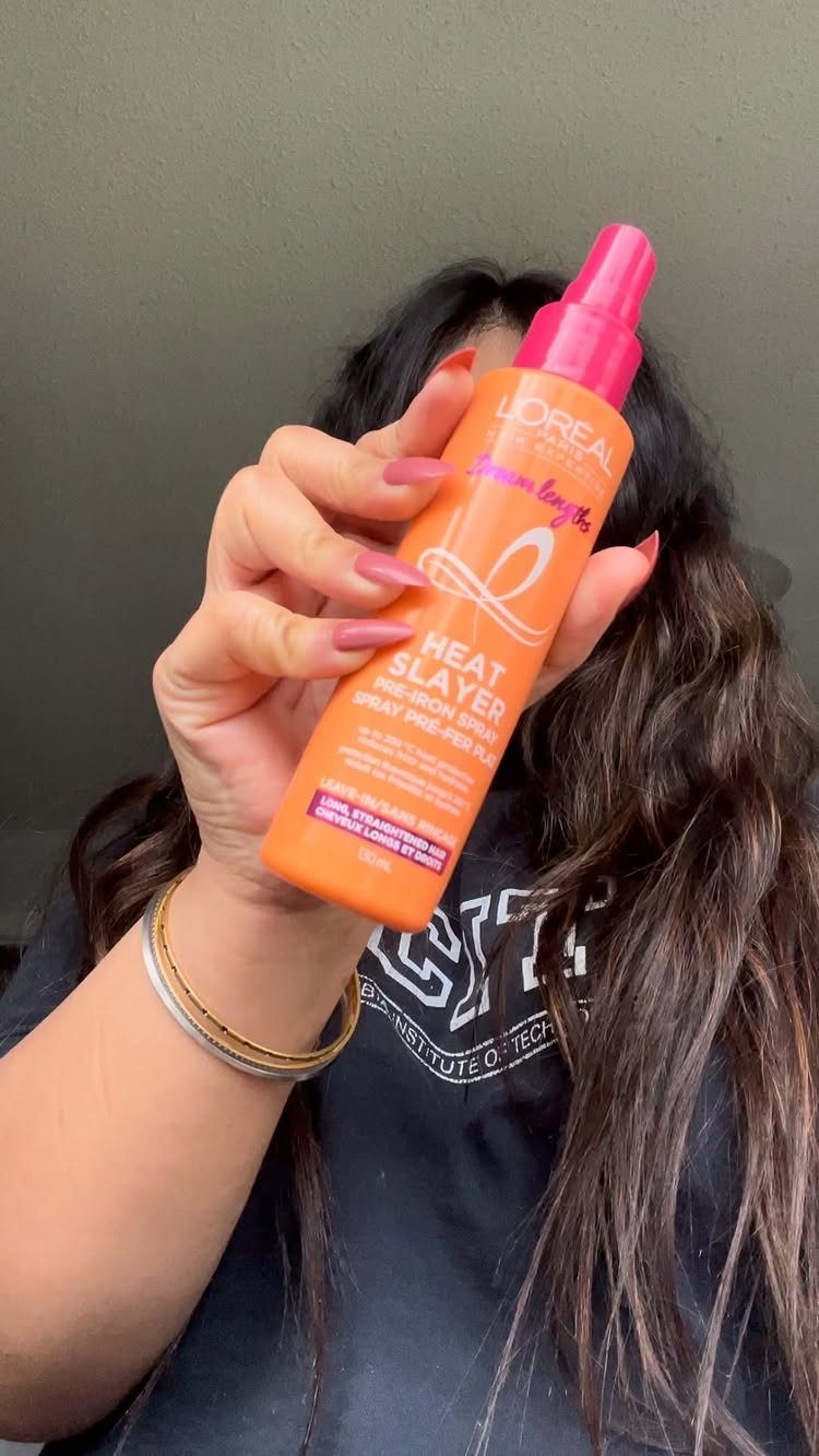 Hand holding an orange L'Oreal Heat Slayer hair spray bottle against grey background