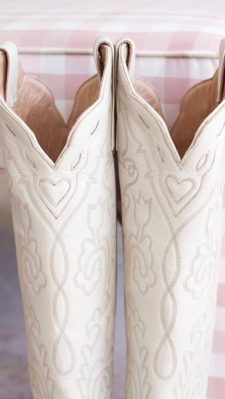 Fall Cowboy Boots for Women: 9 Ideas to Wear Now 7 White leather cowboy boots with decorative stitching against a pink checkered background.