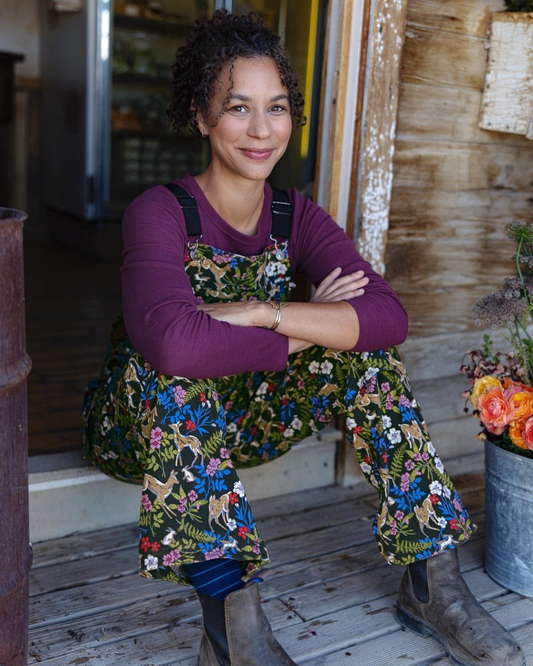 Gillian Practical Magic Outfits: 11 Ideas to Wear Now 4 Woman wearing botanical print overalls and a purple shirt sitting on a porch.