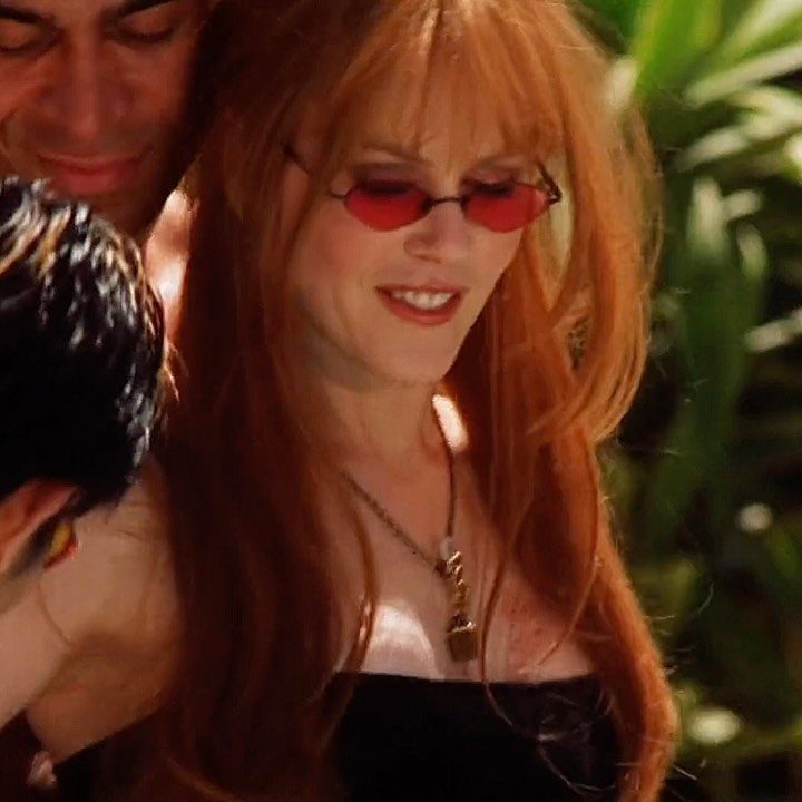 Gillian Practical Magic Outfits: 11 Ideas to Wear Now 7 Woman with red hair wearing red sunglasses and a black strapless top outdoors.