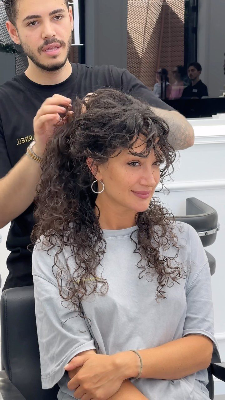 Woman with long dark curly hair and curly bangs being styled by a hairdresser.