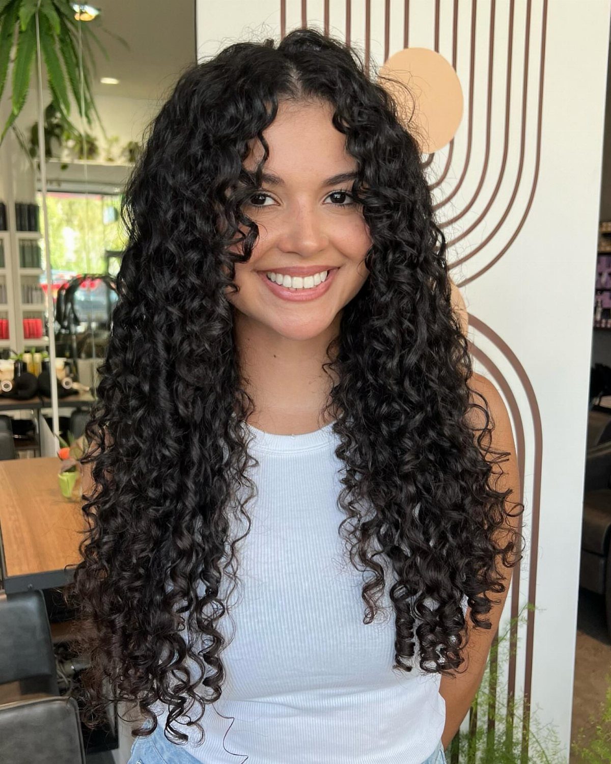 Woman with long, voluminous dark curly hair and bangs smiling in a bright room.