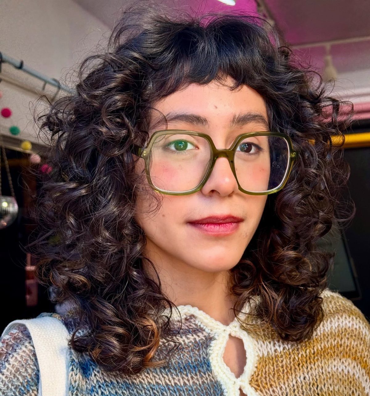 14 Hairstyles with Curly Bangs for Every Length and Curl 9 Person with dark curly hair, short curly baby bangs, and olive-green glasses.