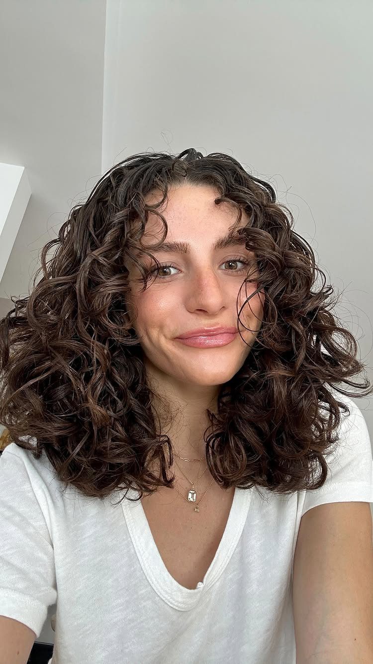 14 Hairstyles with Curly Bangs for Every Length and Curl 3 Woman with voluminous dark brown curly hair and face-framing curly bangs wearing white shirt.