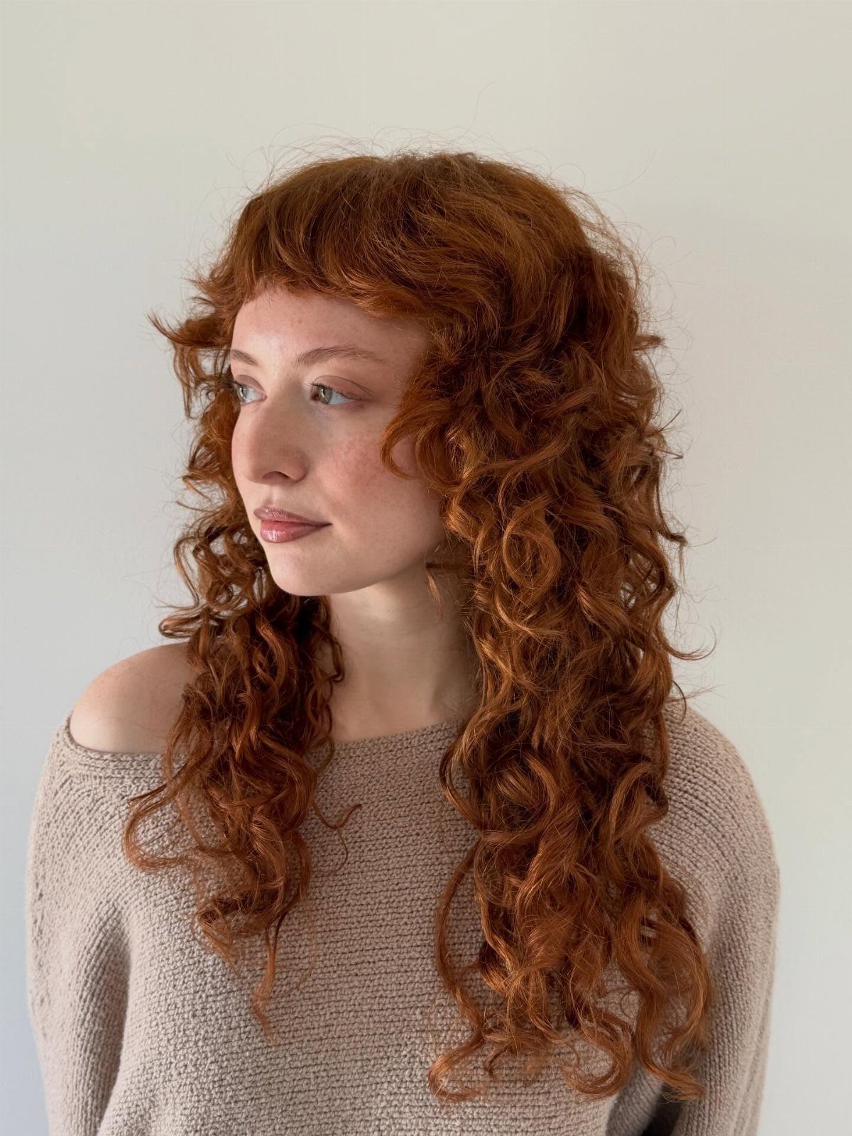 14 Hairstyles with Curly Bangs for Every Length and Curl 11 Woman with long curly red hair and curly bangs wearing a beige sweater
