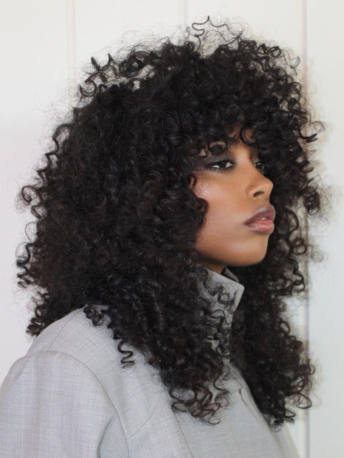 14 Hairstyles with Curly Bangs for Every Length and Curl 13 Woman with voluminous dark curly hair and curly bangs wearing a grey blazer.
