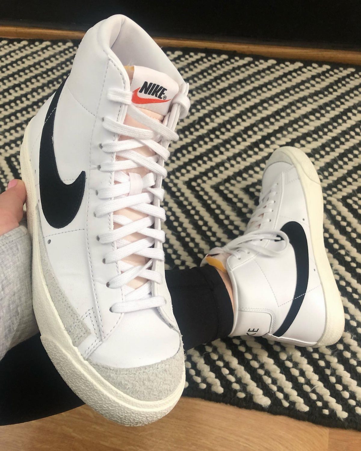 White Nike Blazer Mid 77 sneakers with black swooshes on a patterned rug.