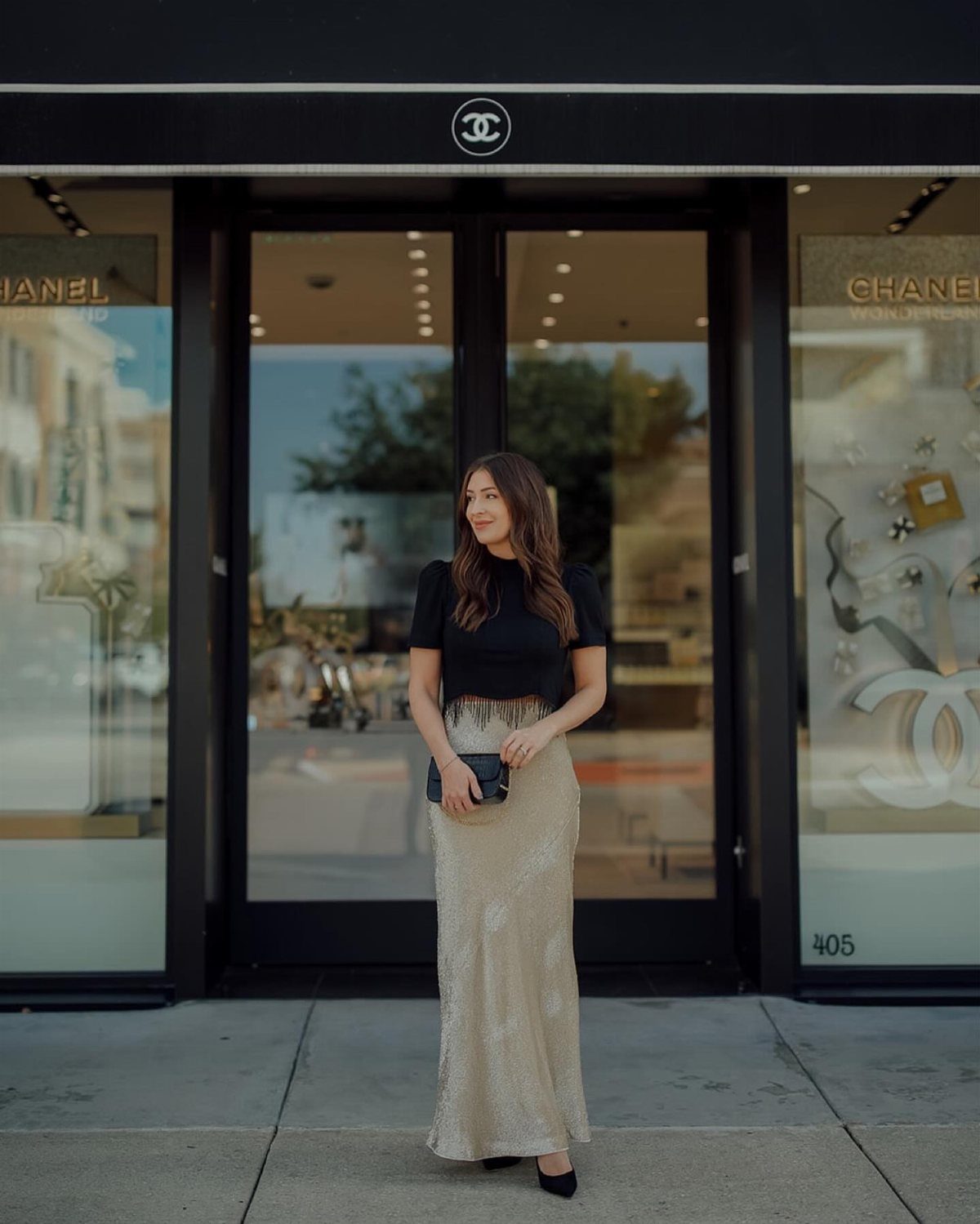 8 Holiday Party Attire for Women to Wear This Season 7 Woman wearing a black top and long gold sequined skirt outside a store.