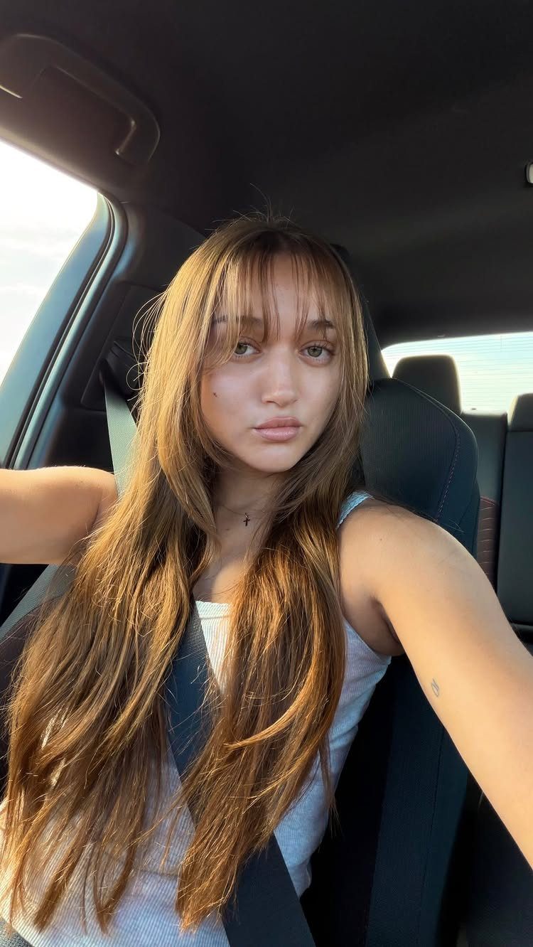 Woman with long brown hair and curtain bangs sitting inside a car.