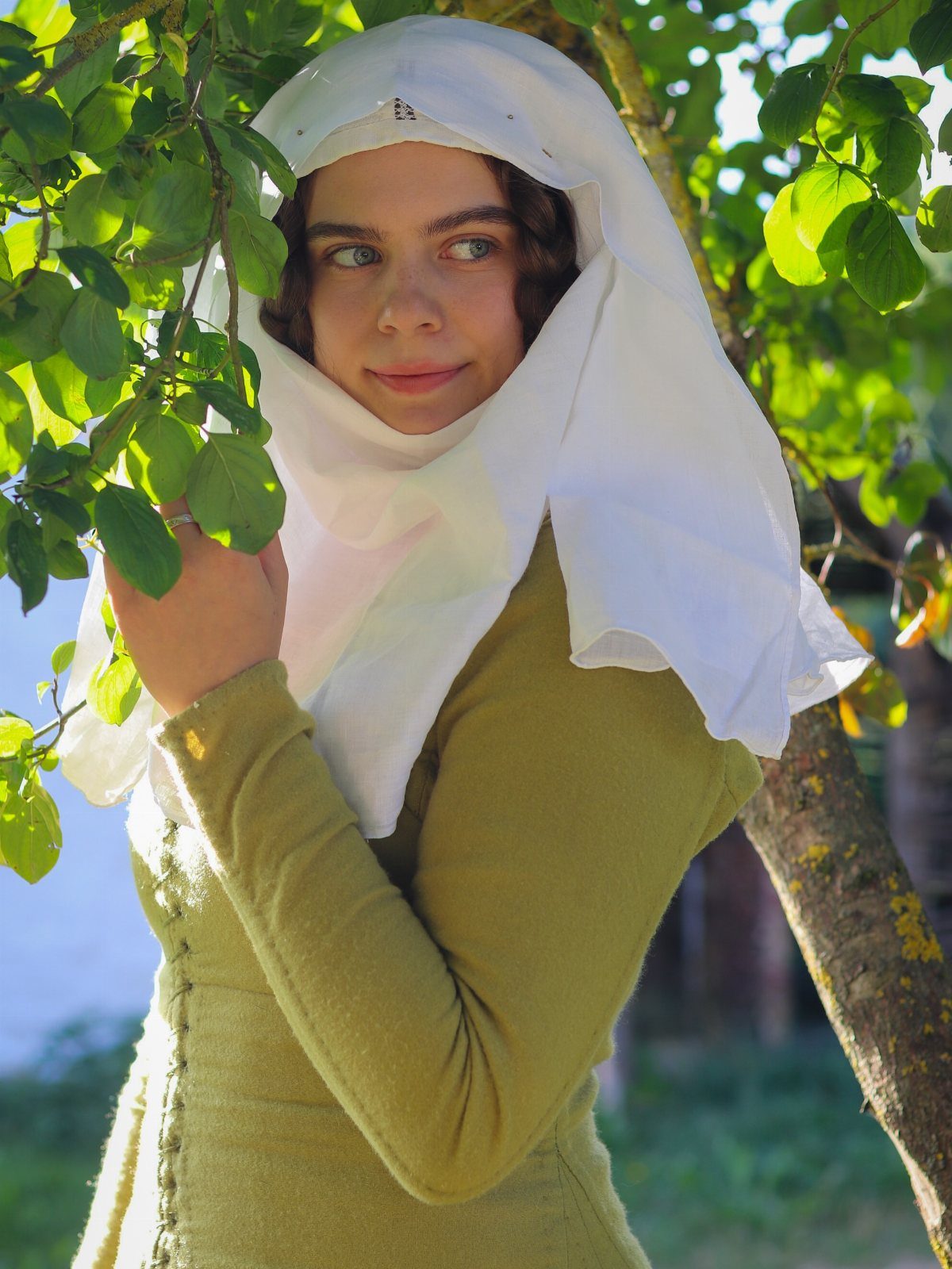 Woman in olive green medieval dress and white head covering among green leaves