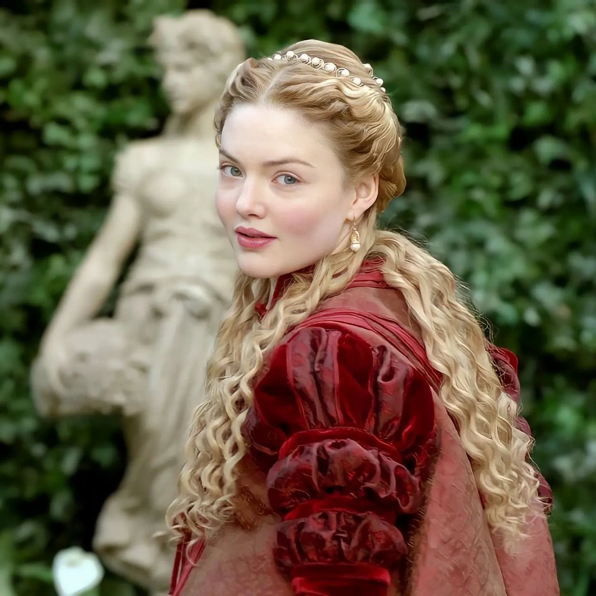 Woman in red velvet medieval gown with pearl headband and curly blonde hair
