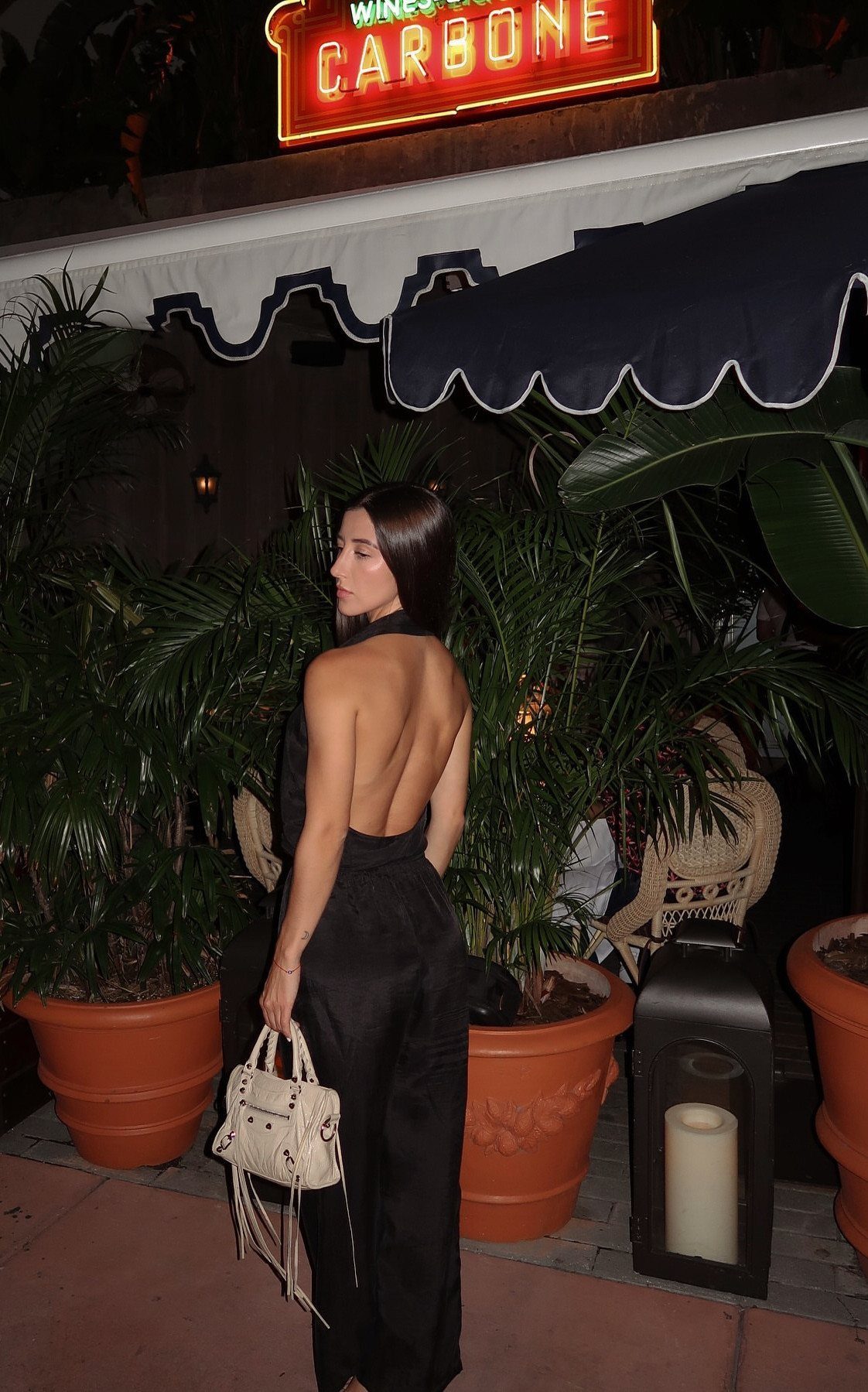 Miami Club Outfits: 8 Ideas for Day and Night 6 Woman in a backless black jumpsuit holding a white bag near tropical plants.
