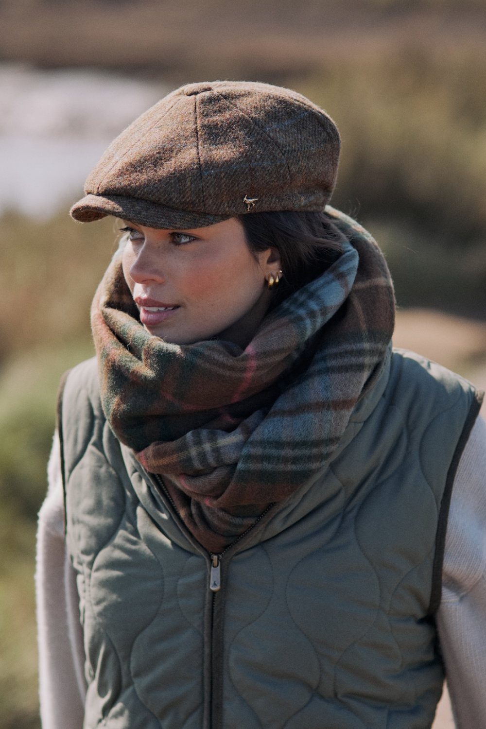 Woman wearing a brown tweed flat cap and a plaid scarf outdoors.
