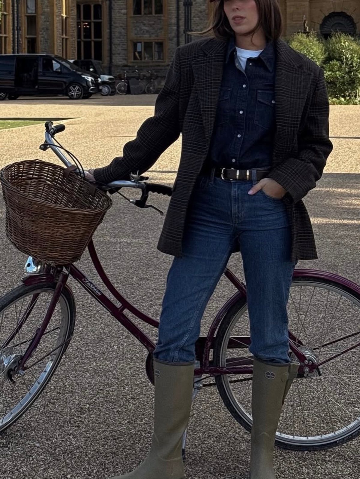 Person in plaid blazer and olive green Wellington boots standing by a bicycle.