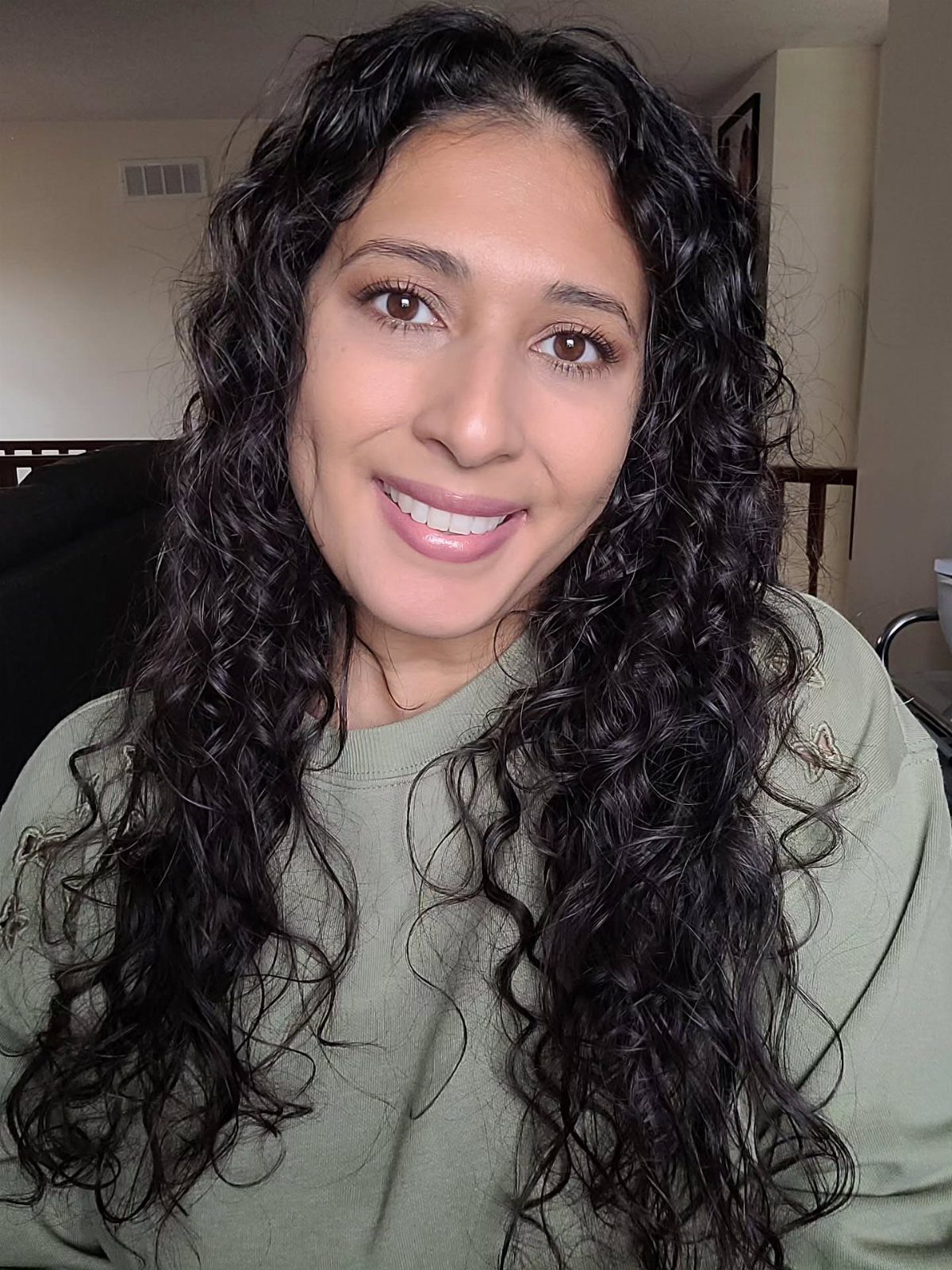 Natural Long Hairstyles: 10 Ideas, From Bantu Knots to Waves 3 Woman with long dark curly hair smiling while wearing an olive green sweatshirt.