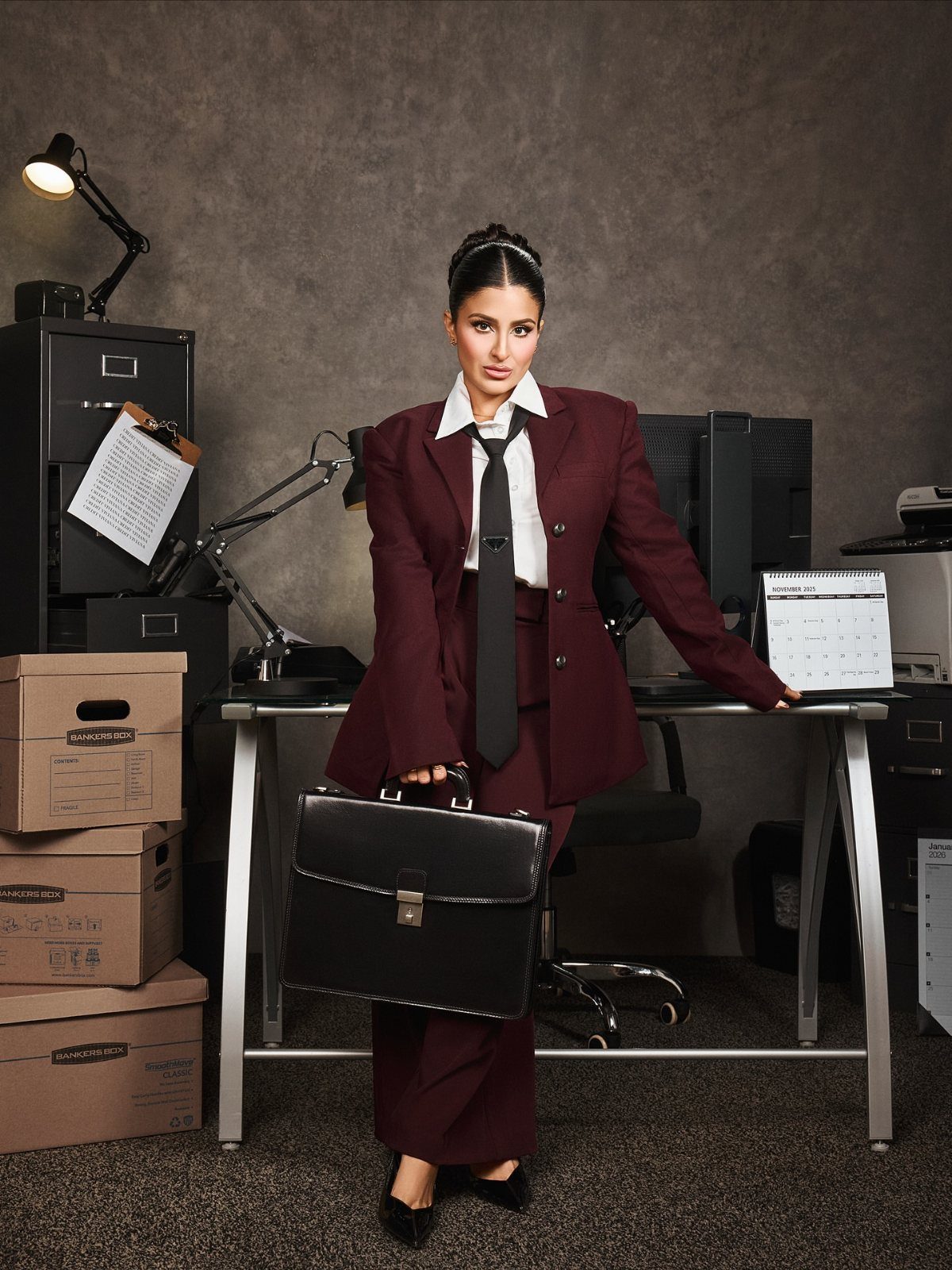 Woman in a burgundy pantsuit and black tie holding a briefcase in an office