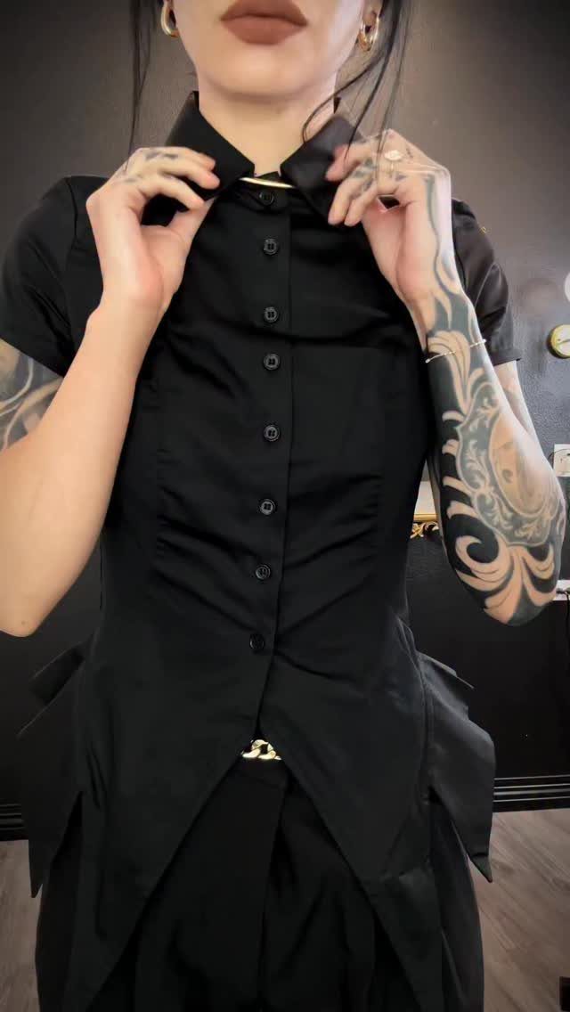 Person with arm tattoos wearing a fitted black button-down shirt and black skirt.
