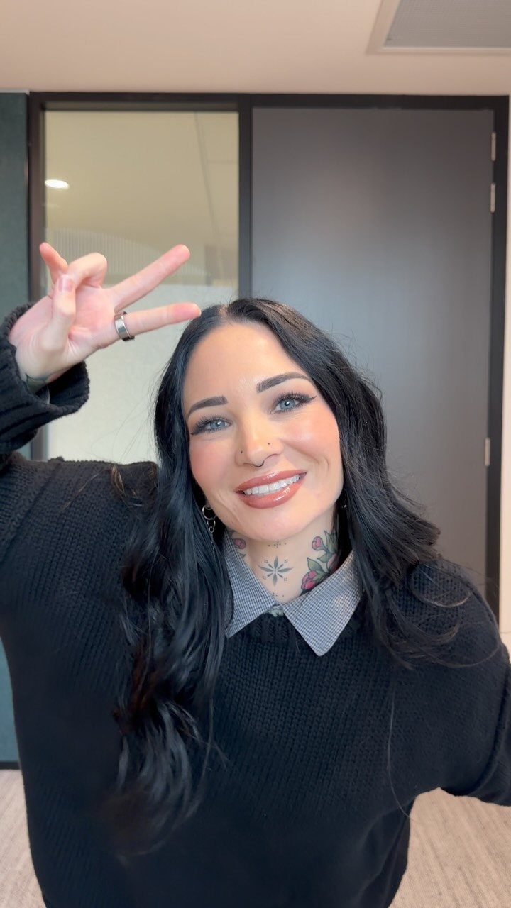 Woman with black hair and tattoos wearing a black sweater and collared shirt.