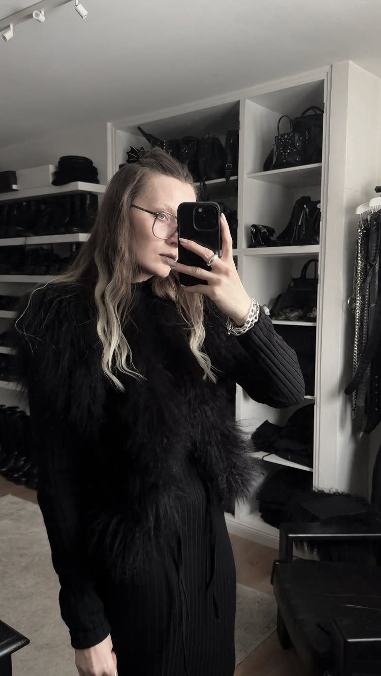 Woman in black ribbed dress and faux-fur vest posing in a white closet.