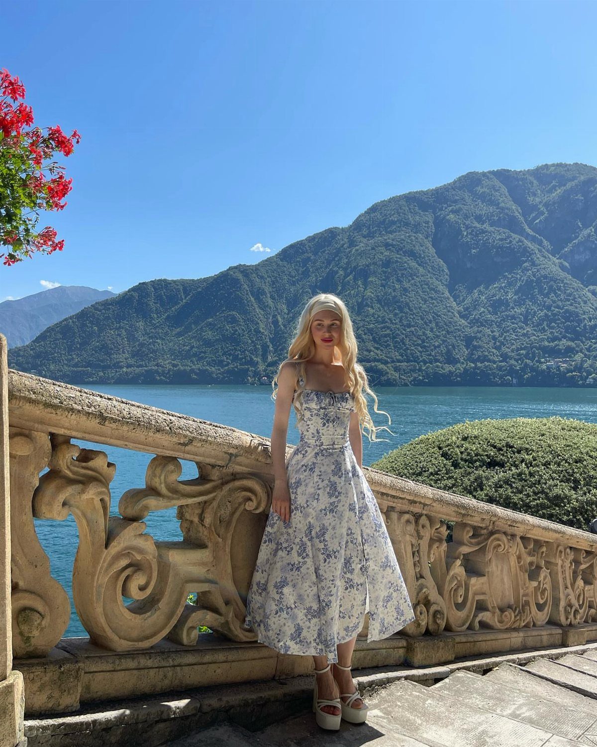Woman in a blue floral vintage dress standing by a stone railing overlooking mountains.