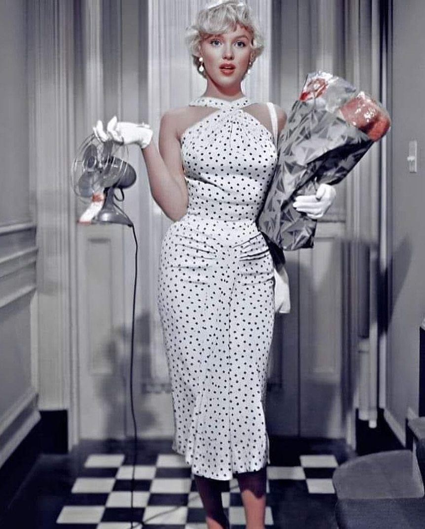 Blonde woman in a white polka dot midi dress holding flowers and a fan.
