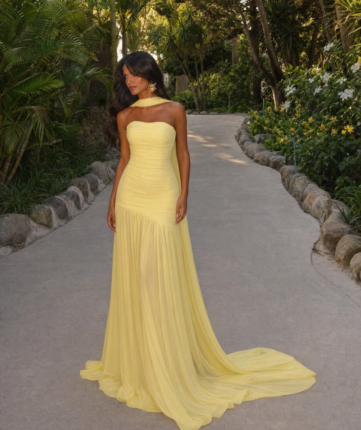 Woman in a pale yellow one-shoulder draped maxi dress on a garden path